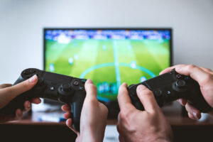 Zwei Hände halten zwei Playstation Controller. Im Hintergrund steht ein Fernseher auf dem das Spiel Fifa läuft.